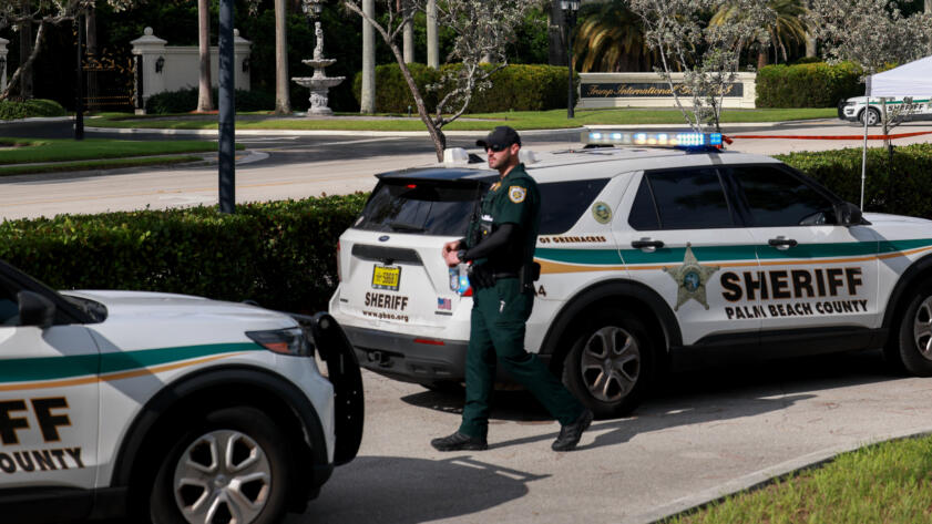 A law enforcement officer in a dark green uniform walks in between two police cars with the Palm Beach County Sheriff’s Office logo on the side. The acrs are parked across the street from the entrance of a golf course.